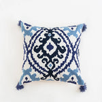 Coussin décoratif aux motifs géométriques bleus et noirs sur fond blanc, avec des franges sur les côtés.