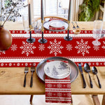 La table est élégamment décorée pour un repas de fête avec des motifs de flocons de neige rouges et blancs sur la nappe, des bougies et des assiettes avec des motifs de paysage enneigé, le tout créant une ambiance chaleureuse et festive pour les convives.