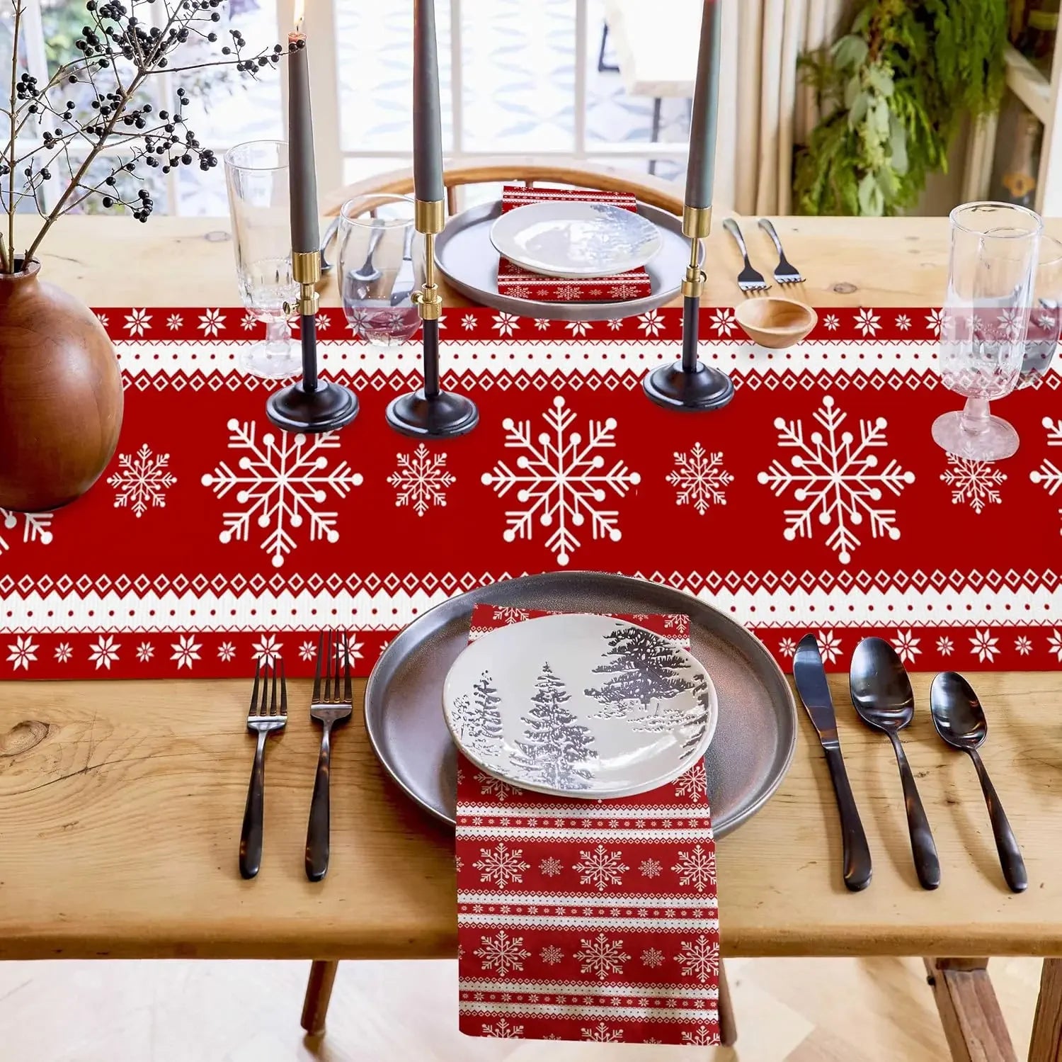 La table est élégamment décorée pour un repas de fête avec des motifs de flocons de neige rouges et blancs sur la nappe, des bougies et des assiettes avec des motifs de paysage enneigé, le tout créant une ambiance chaleureuse et festive pour les convives.