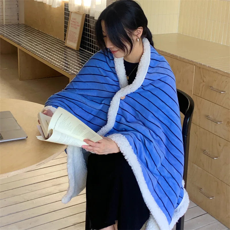 Une femme assise dans une pièce, lisant un livre tout en étant enveloppée dans une couverture rayée bleue et blanche.