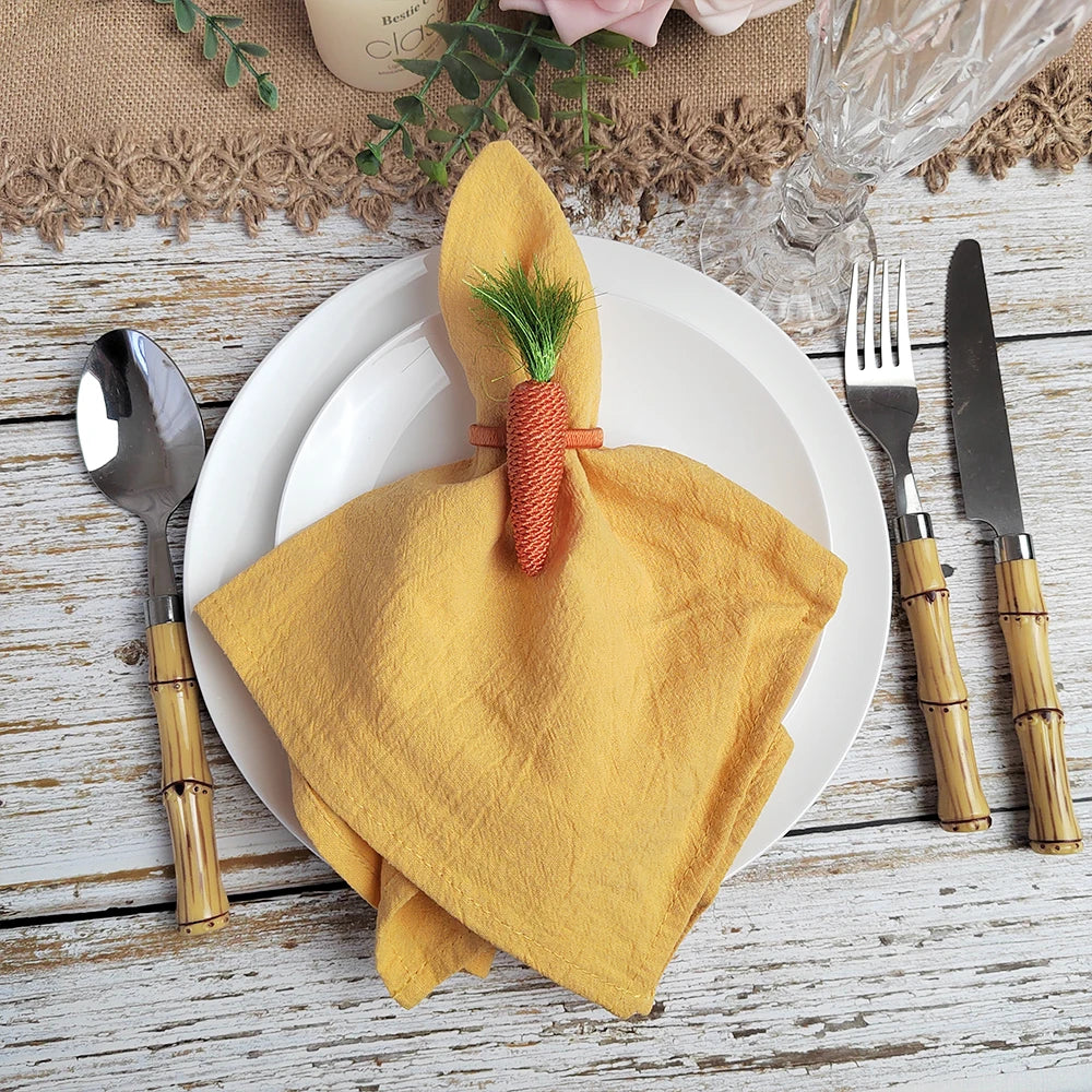 Sur une assiette blanche, une serviette jaune est enroulée et attachée avec un élément décoratif représentant une carotte. L'assiette est posée sur une table en bois rustique, accompagnée de couverts en bois.