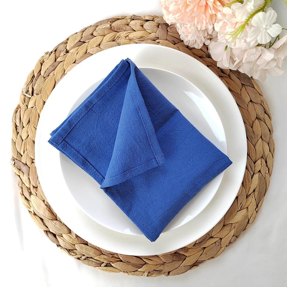Une serviette en tissu bleu foncé est posée sur une assiette blanche, entourée d'un napperon en tresses de fibres végétales et de fleurs.