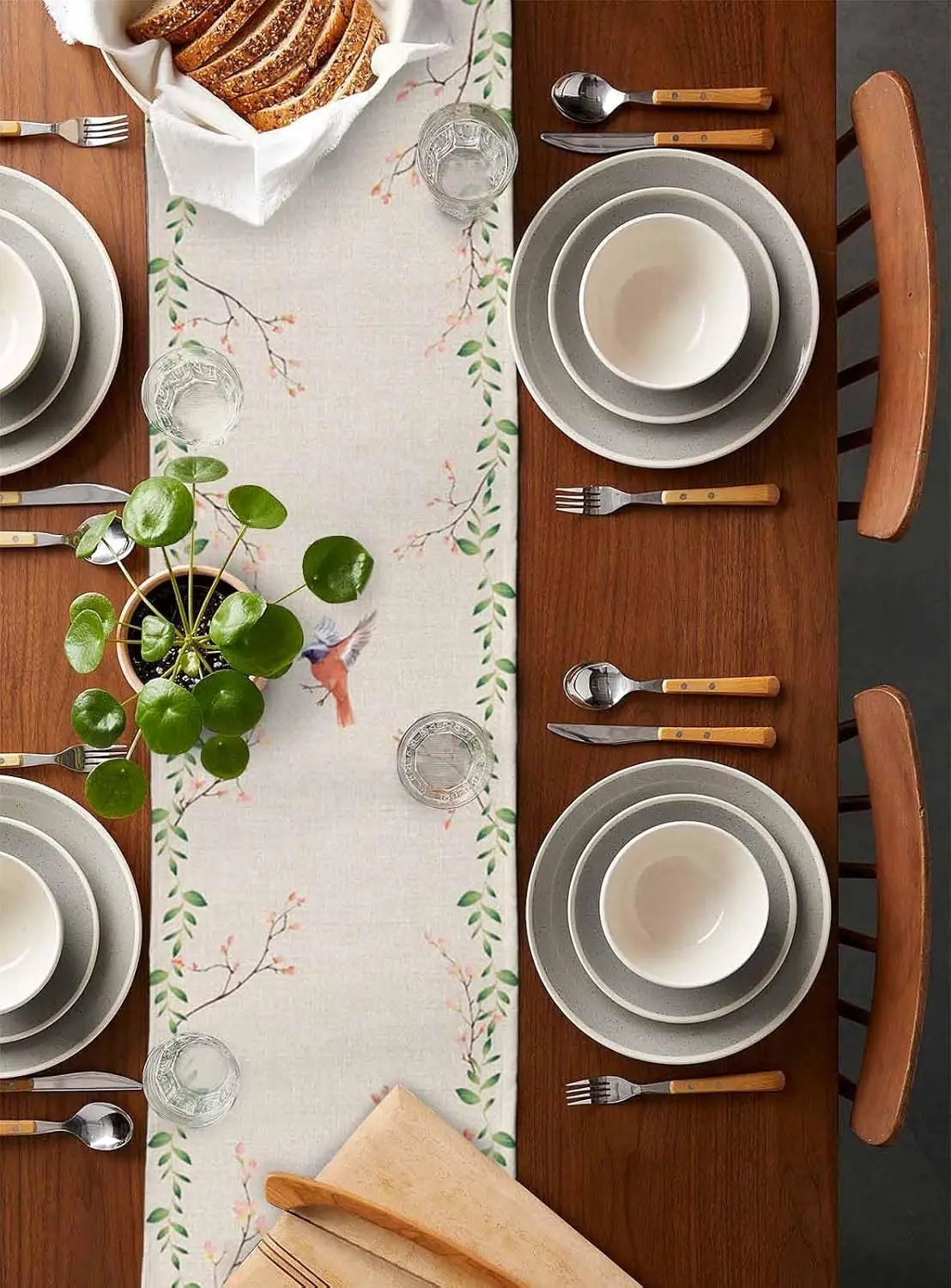 Une table de salle à manger joliment dressée avec de la vaisselle grise, des couverts en bois et une jolie nappe à motifs floraux, accompagnés d'un petit pot de plante verte, créant une ambiance chaleureuse et élégante.