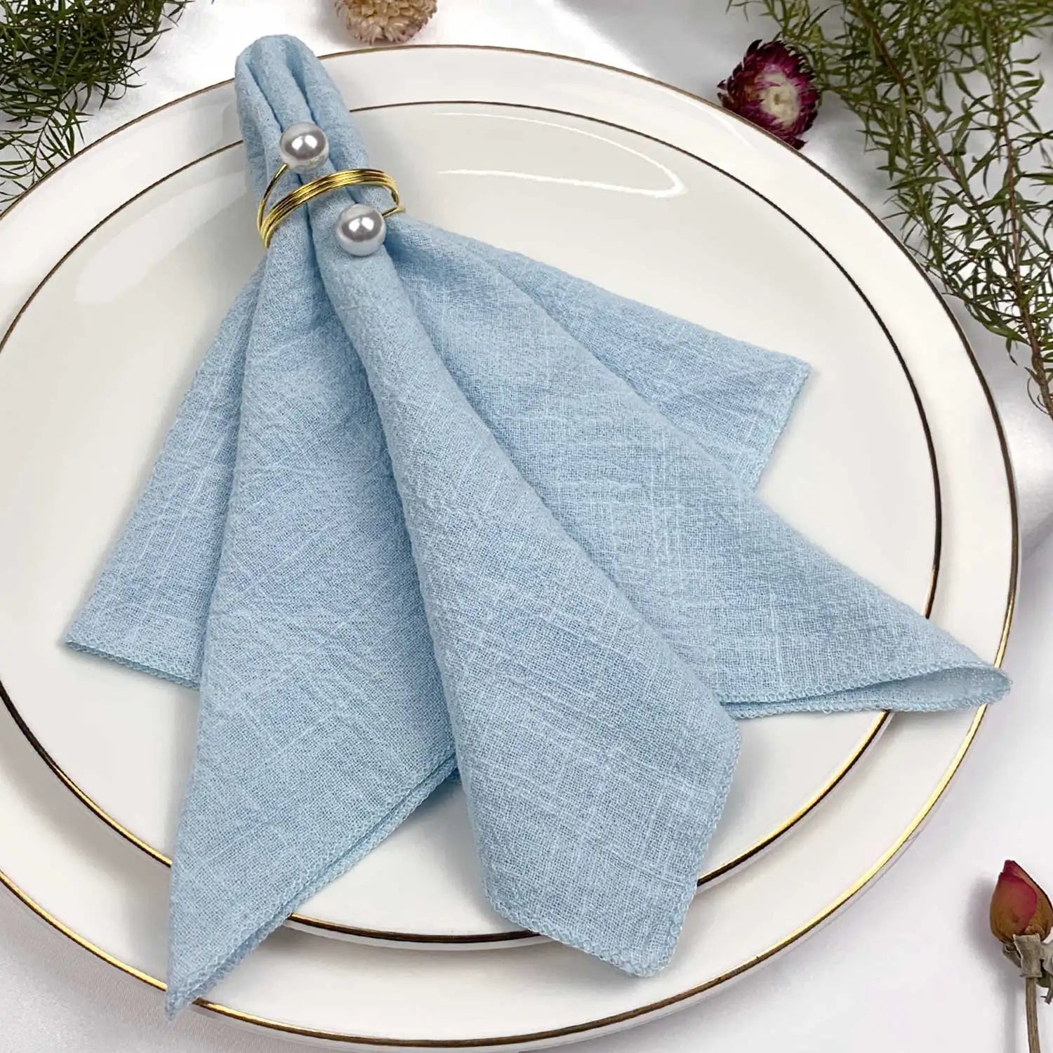 Serviette de table grise décorée de perles dorées, sur une assiette blanche avec un cadre doré et des accessoires floraux.