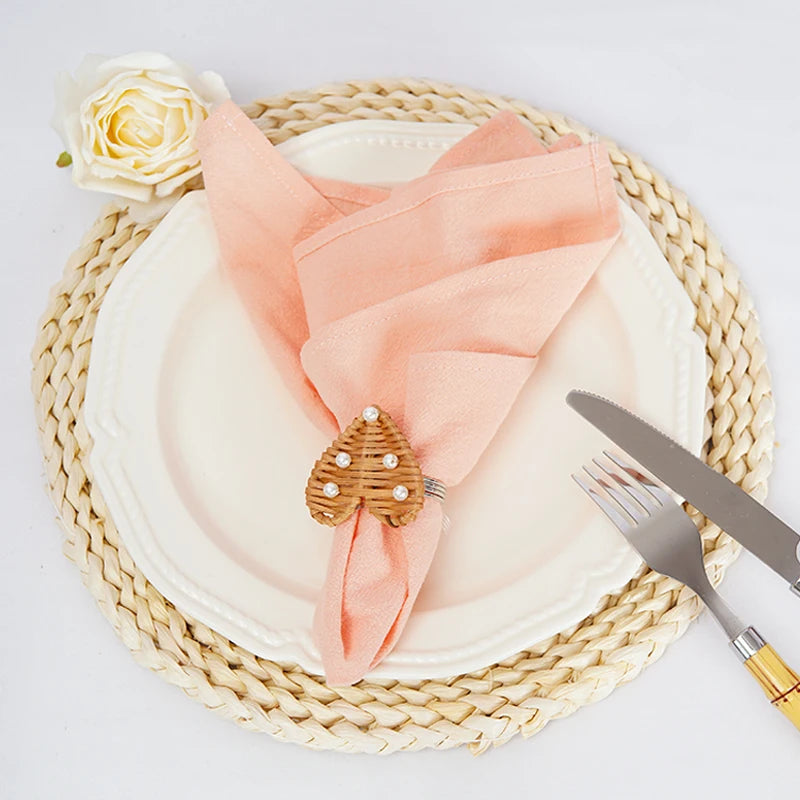 Une décoration élégante pour la table, avec un napperon en fibres tressées, une assiette blanche, une serviette rose pâle et un accessoire en forme de pomme de pin décoré de strass.