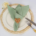 Un élégant arrangement de table avec une serviette de table verte pliée et décorée d'un cœur en rotin, accompagné d'une fourchette et d'un couteau, sur un napperon tressé beige.