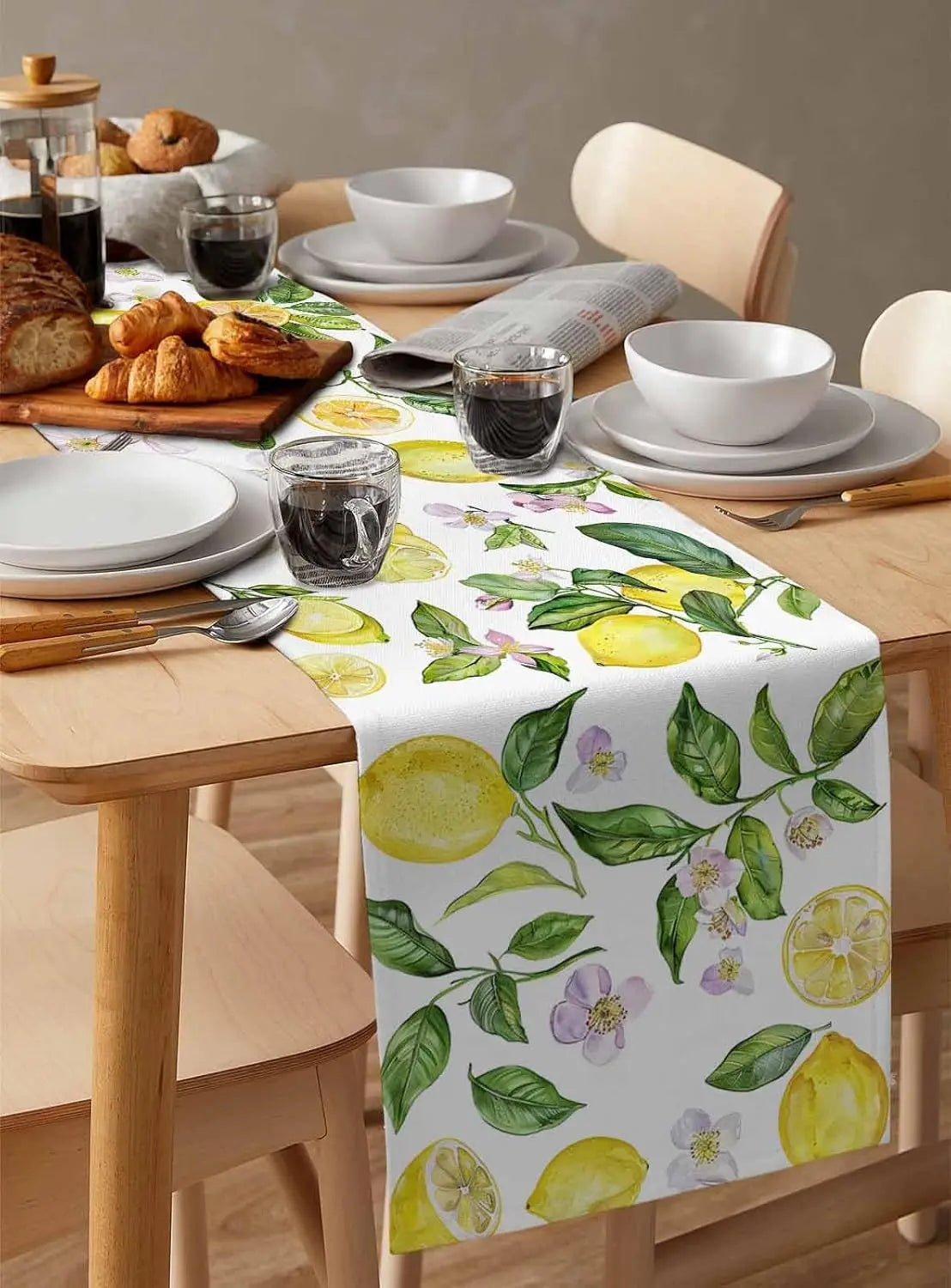 La table de petit-déjeuner est garnie de produits de boulangerie, de vaisselle blanche et de verres, le tout posés sur une nappe imprimée de motifs végétaux aux couleurs fraîches et ensoleillées.