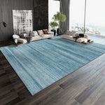 Une pièce de vie moderne avec un grand tapis bleu vert, des canapés confortables et des plantes verdoyantes offrant une vue panoramique sur un paysage extérieur.