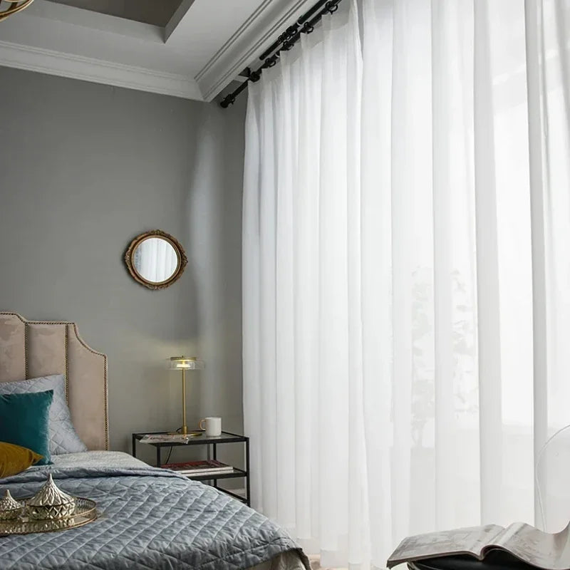 Chambre élégante avec voilages blancs laissant passer une douce lumière naturelle, créant une atmosphère apaisante. Décoration raffinée avec un lit confortable, oreillers de couleurs variées, et une lampe moderne sur une table de chevet noire. Design intérieur moderne parfait pour un lieu de repos et relaxation.