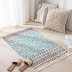 Tapis décoratif de style boho avec motifs géométriques et franges, posé sur le sol à côté de coussins décoratifs et de sandales.