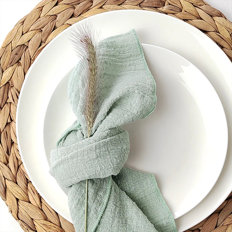Assiette blanche avec serviette de table verte enroulée sur un dessous de plat tressé en fibres végétales, disposés sur une surface de table blanche.