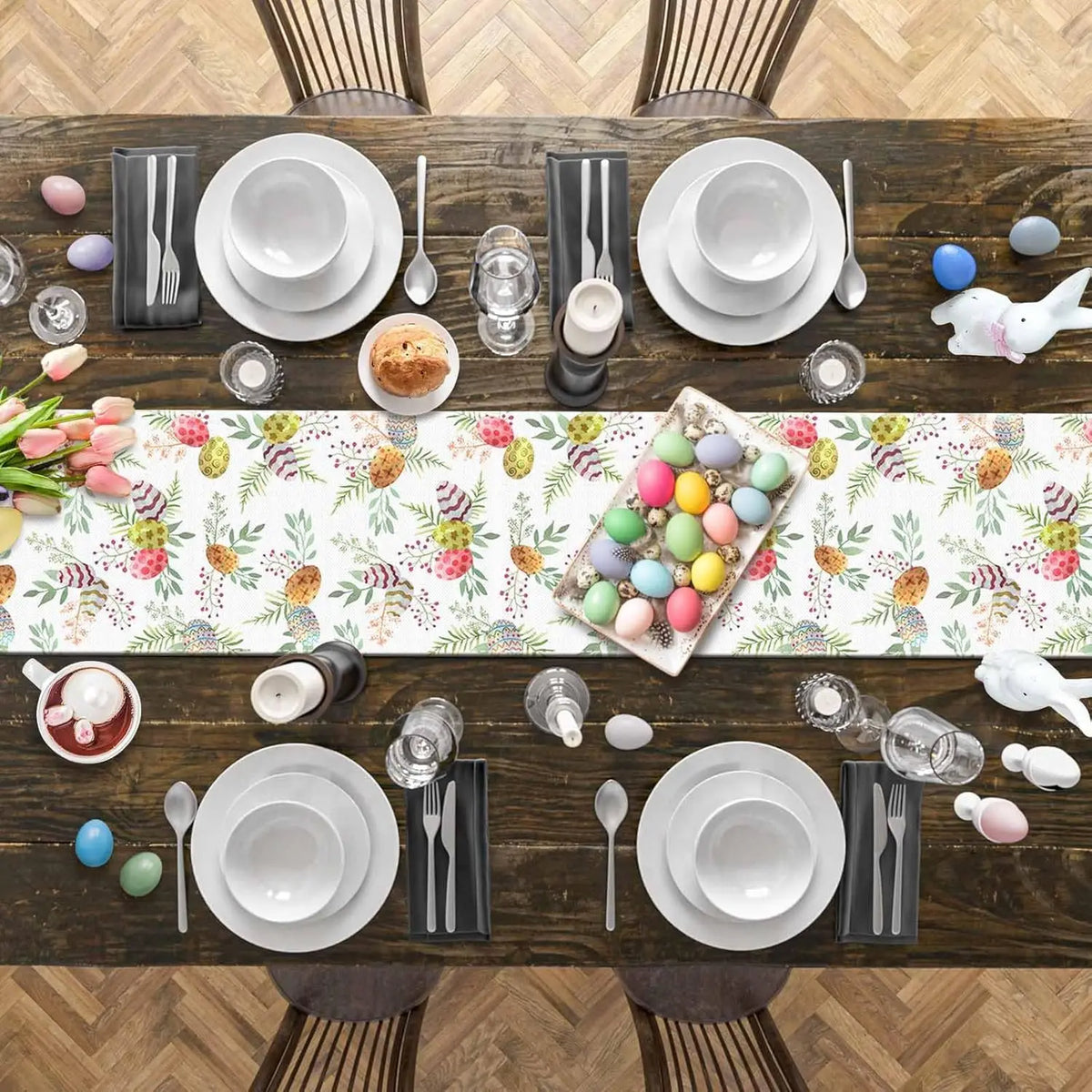 Cette image montre une table en bois avec une nappe de motifs printaniers, décorée pour un repas de Pâques avec de la vaisselle blanche, des couverts, des bougies, des tulipes et des œufs colorés.