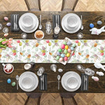 Cette image montre une table en bois avec une nappe de motifs printaniers, décorée pour un repas de Pâques avec de la vaisselle blanche, des couverts, des bougies, des tulipes et des œufs colorés.