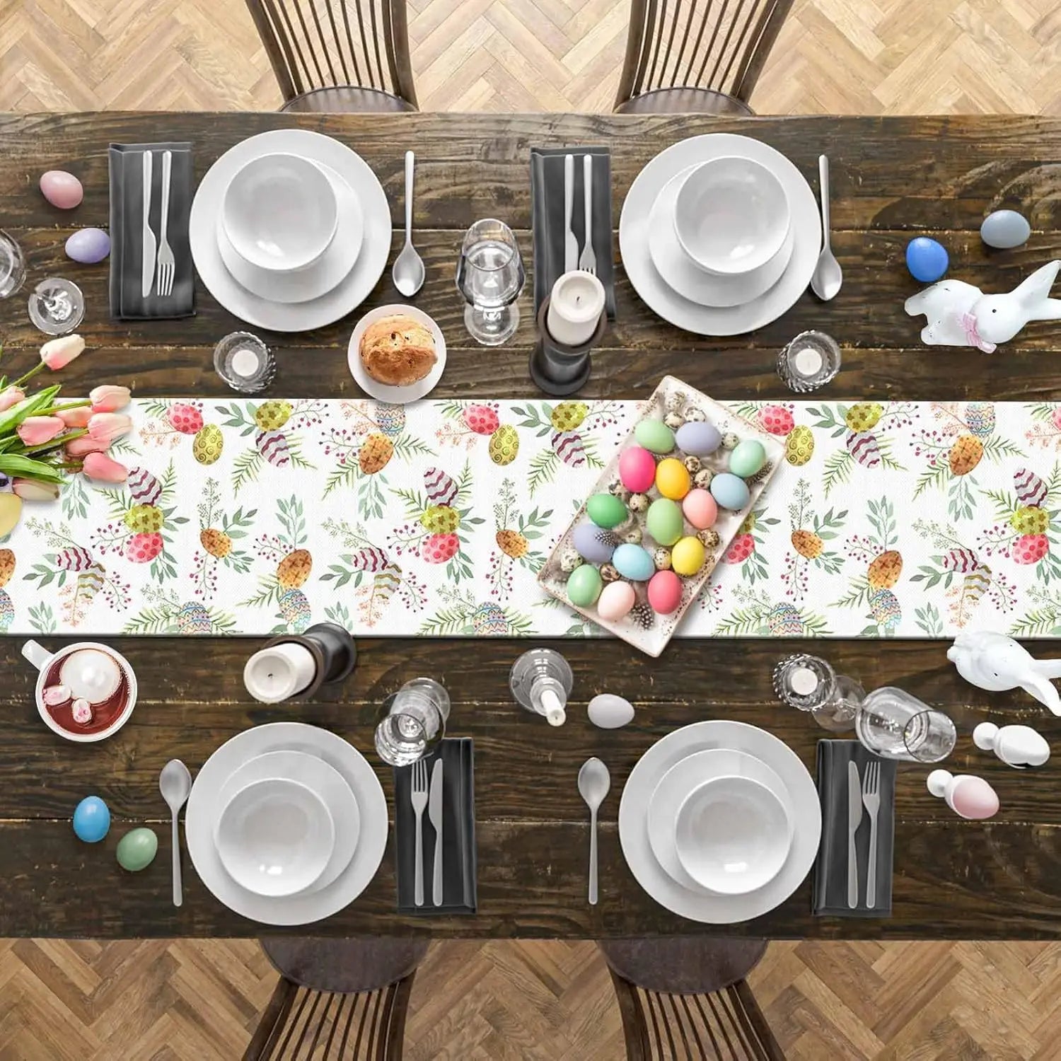 Cette image montre une table en bois avec une nappe de motifs printaniers, décorée pour un repas de Pâques avec de la vaisselle blanche, des couverts, des bougies, des tulipes et des œufs colorés.