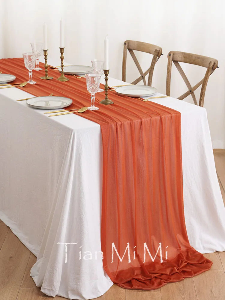 Magnifique table de salle à manger élégamment dressée avec une nappe blanche, un chemin de table orange vif, des verres de cristal et des bougeoirs dorés, le tout sur un parquet en bois clair.