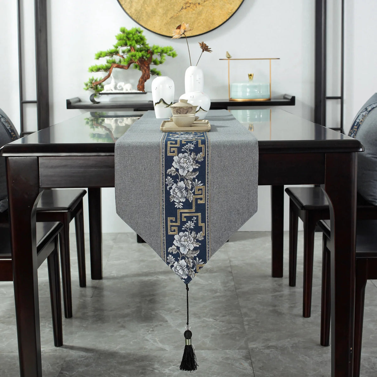 Une table à manger en bois sombre avec un chemin de table gris brodé de motifs floraux et de bordures géométriques ornant élégamment le centre de la pièce.