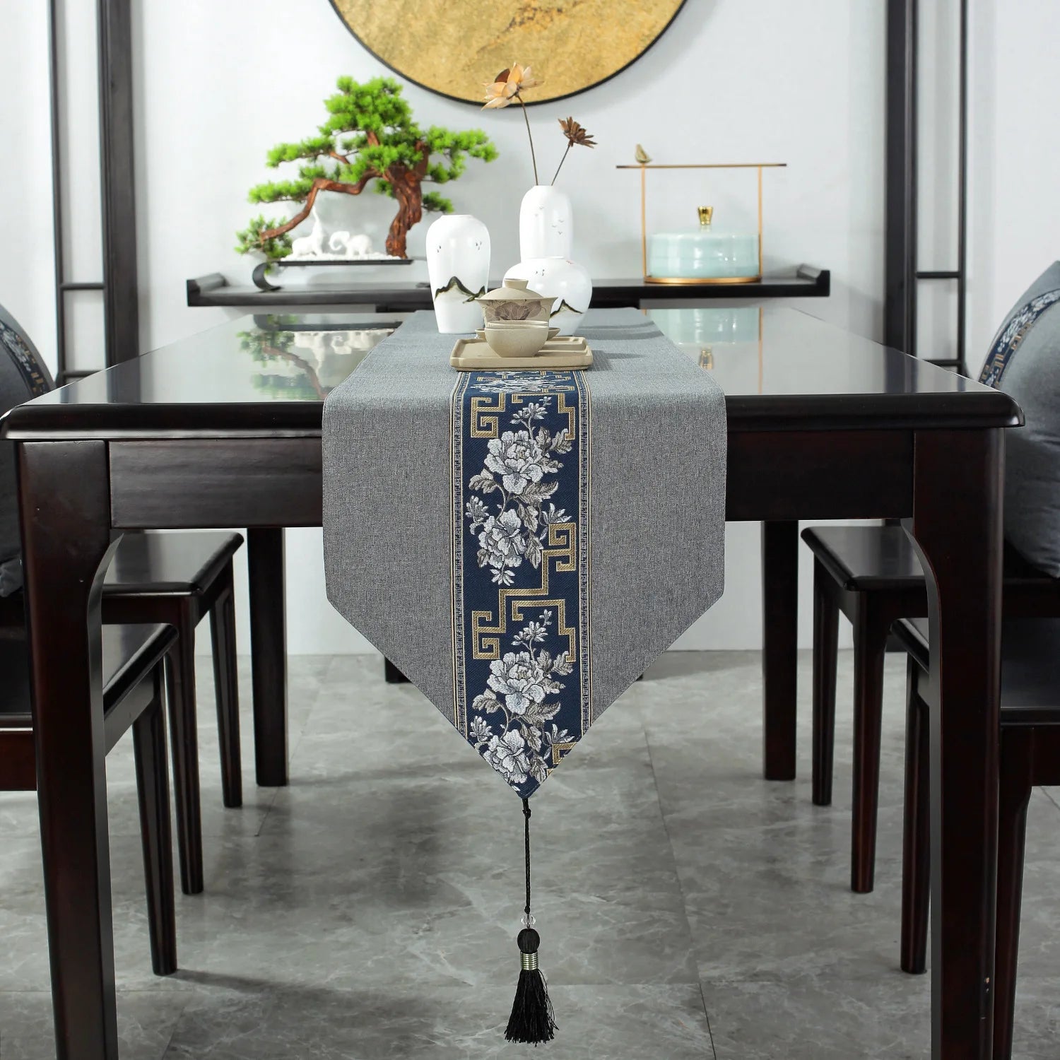 Une table à manger en bois sombre avec un chemin de table gris brodé de motifs floraux et de bordures géométriques ornant élégamment le centre de la pièce.
