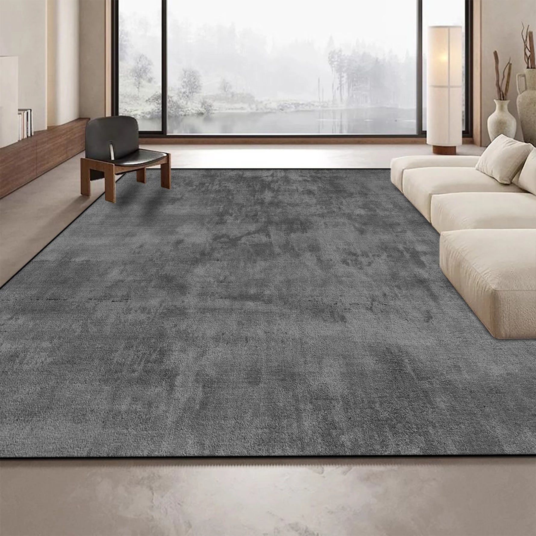 Un tapis gris foncé recouvre le sol d'un salon spacieux avec de grandes fenêtres offrant une vue sur un paysage forestier enneigé.