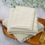 Des serviettes de table en lin beige sont empilées sur un plateau en bois, contre un fond flou de verdure, créant une atmosphère apaisante et naturelle.