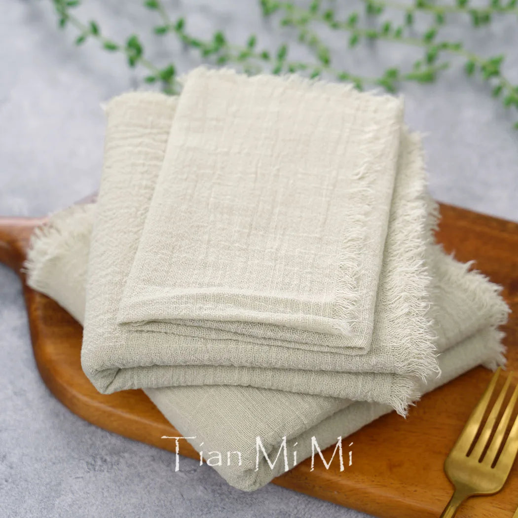 Des serviettes de table en lin beige sont empilées sur un plateau en bois, contre un fond flou de verdure, créant une atmosphère apaisante et naturelle.