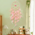 Un capteur de rêves rose suspendu devant une fenêtre, avec des plumes douces et légères, créant une atmosphère paisible et féminine dans cette pièce décorée avec soin.
