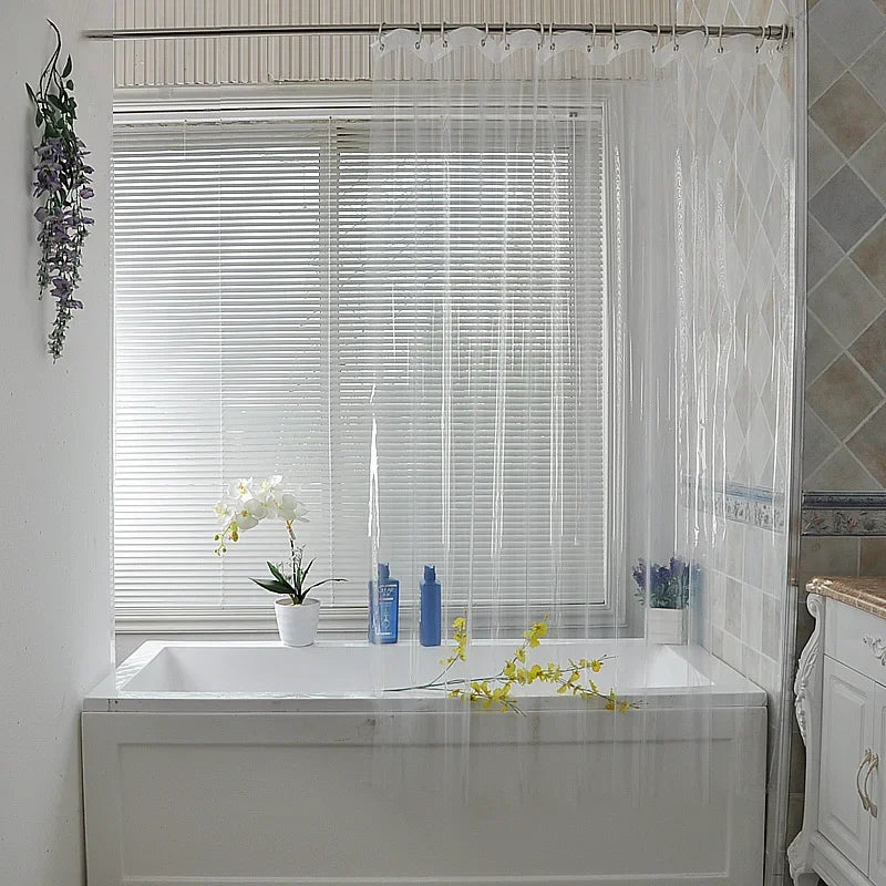 Rideau de douche transparent en vinyle suspendu sur une tringle, entourant une baignoire blanche près d'une grande fenêtre avec stores. Salle de bain lumineuse avec orchidées et carrelage mural.