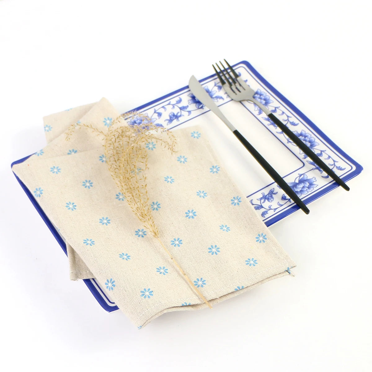 Une serviette à motifs floraux bleus et blancs posée sur une assiette ornée est accompagnée d'une fourchette et d'un couteau sur un fond blanc.