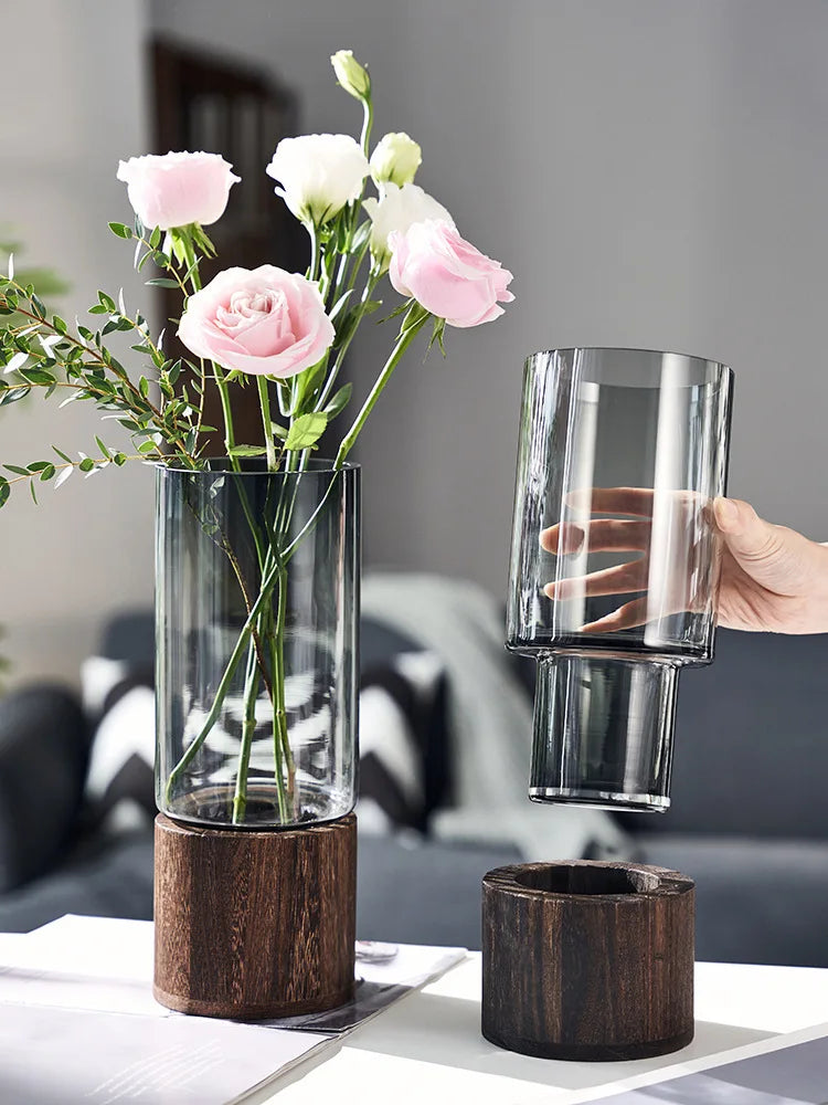 Un bouquet de roses roses délicates dans un vase en verre sur un support en bois, avec un verre en cristal à proximité.