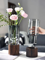 Un bouquet de roses roses délicates dans un vase en verre sur un support en bois, avec un verre en cristal à proximité.