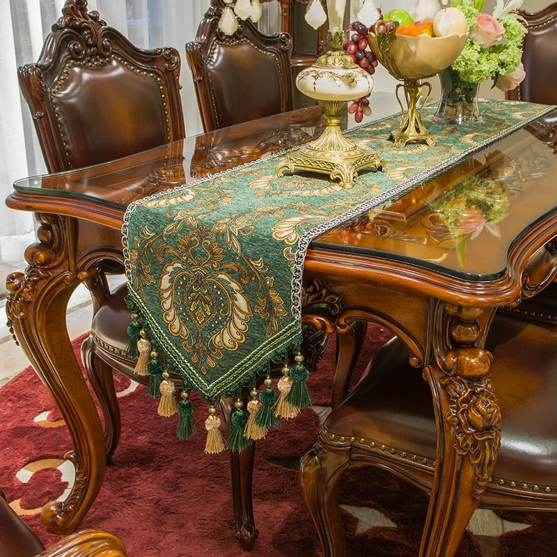 Cette image montre une élégante table en bois foncé avec des chaises ornées, recouverte d'un chemin de table vert et or à motifs floraux et franges assorties, soulignant le style luxueux et raffiné de cette pièce d'ameublement.