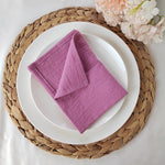 Une serviette de table en lin de couleur mauve sur une assiette blanche posée sur un dessous de plat en tresse de fibres naturelles, avec des fleurs de pivoine en arrière-plan.
