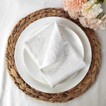 Sur une table blanche, une assiette en porcelaine blanche est posée sur un set de table en fibres tressées. Un linge de table blanc est délicatement plié sur l'assiette, avec un bouquet de fleurs roses et blanches à côté.