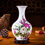 Un vase en céramique blanche décoré de fleurs roses, blanches et vertes sur un fond sombre, avec une monture en bois foncé.
