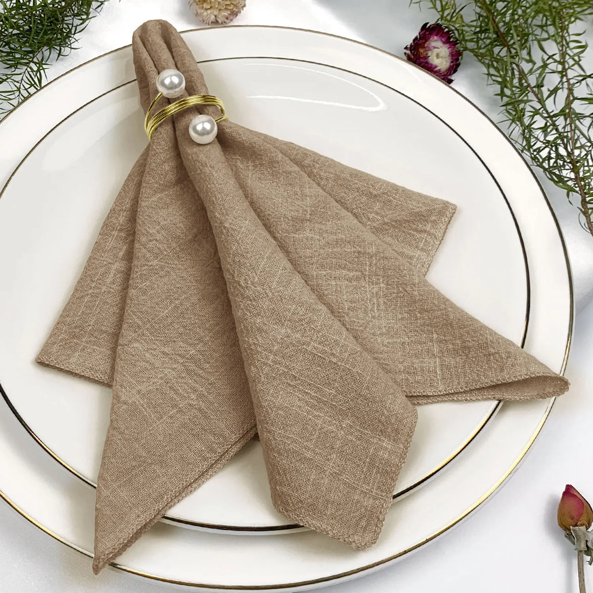 Une belle serviette de table beige ornée de perles dorées, disposée sur une assiette blanche avec des fleurs.