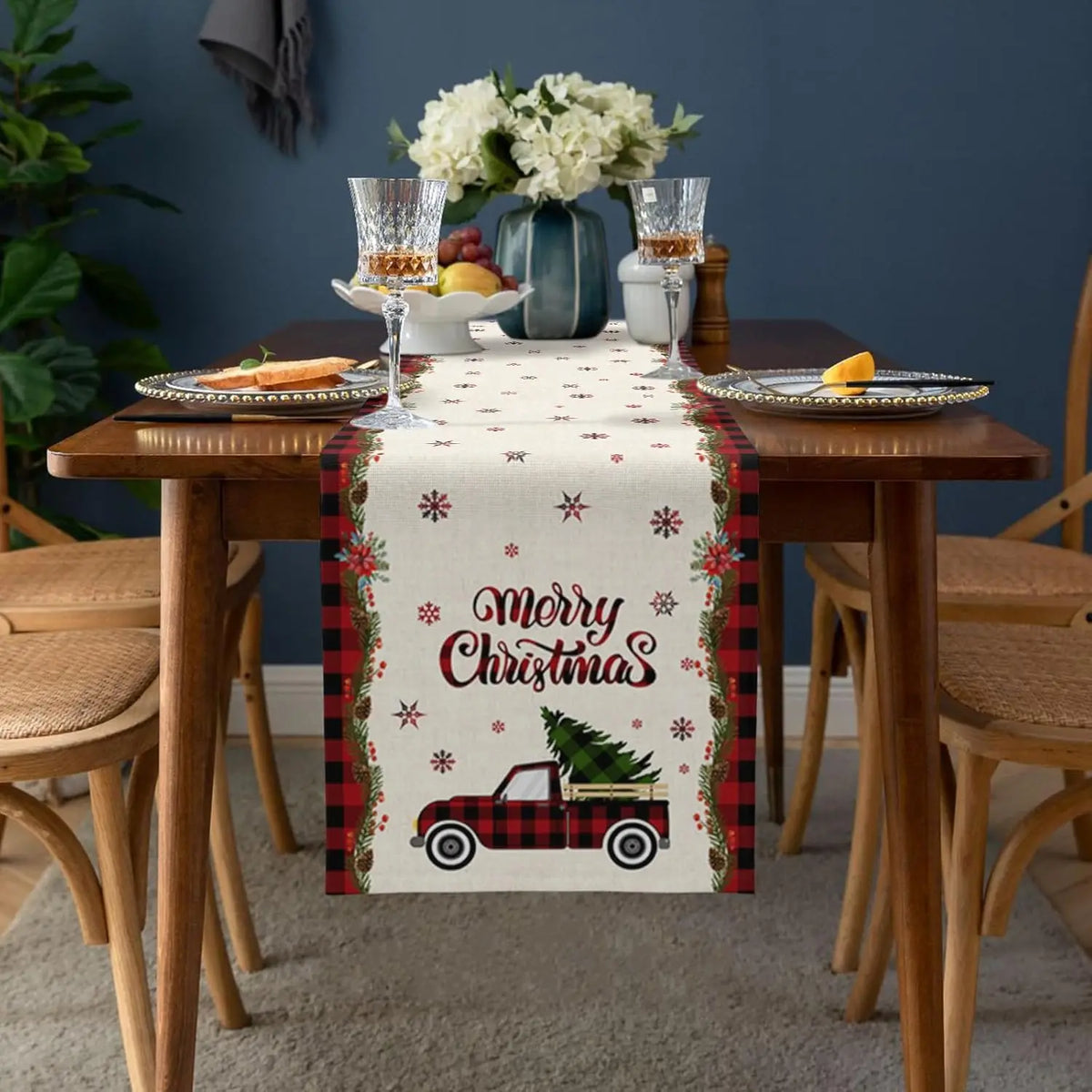 Une table de salle à manger joliment décorée pour Noël, avec un chemin de table à motifs de camion et de flocons de neige, accompagnée de vases de fleurs et de verres à vin.
