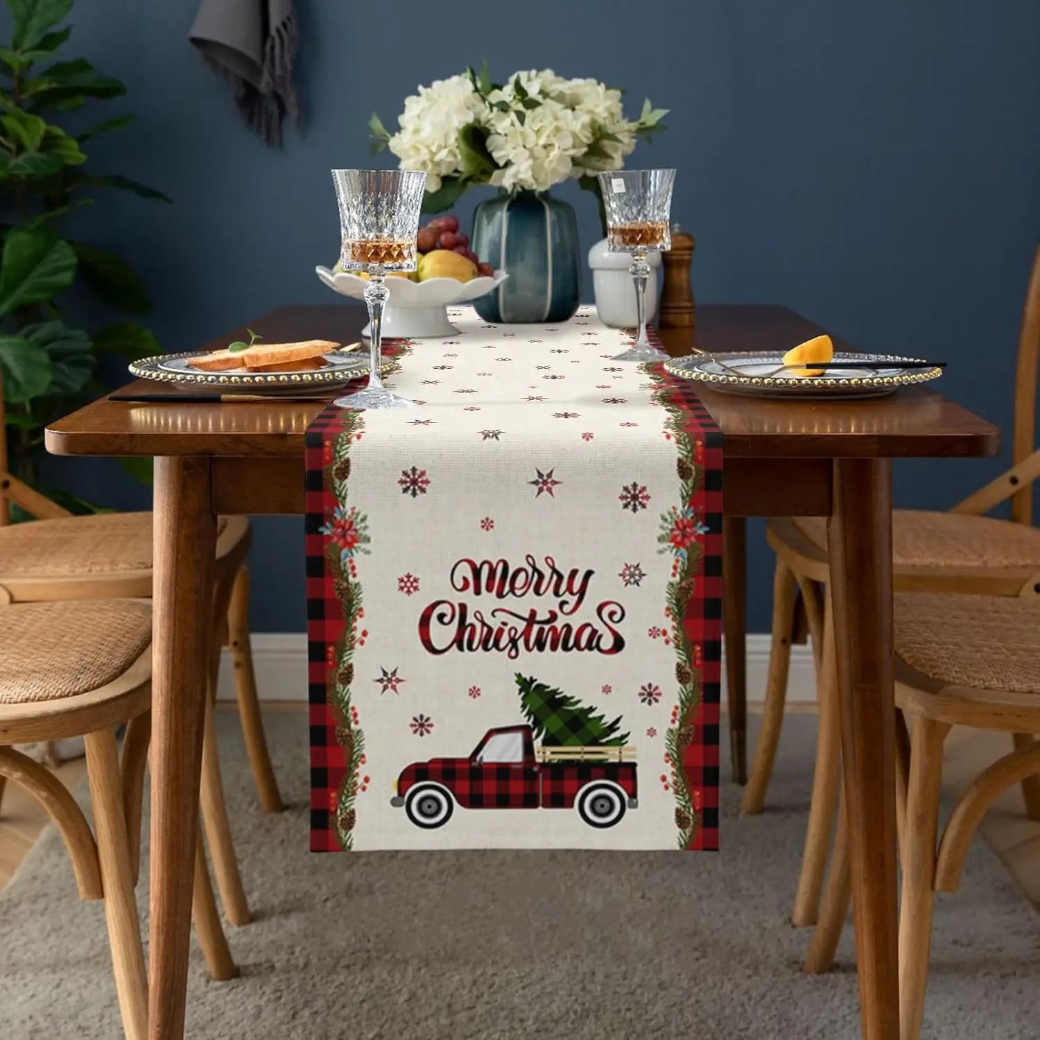 Une table de salle à manger joliment décorée pour Noël, avec un chemin de table à motifs de camion et de flocons de neige, accompagnée de vases de fleurs et de verres à vin.