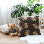 Un coussin décoratif avec des motifs de fleurs et un ours en peluche sur une table en bois, entourés de plantes vertes.