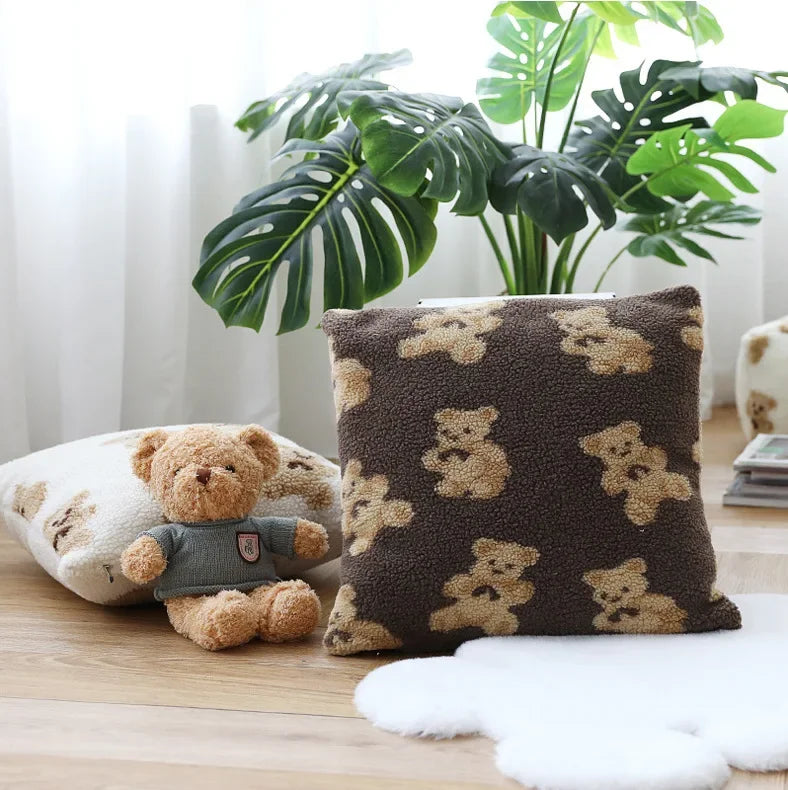 Un coussin décoratif avec des motifs de fleurs et un ours en peluche sur une table en bois, entourés de plantes vertes.
