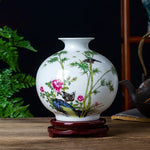 Un vase en porcelaine blanche décoré d'un motif de bambous, de fleurs et d'oiseaux colorés, disposé sur un présentoir en bois.