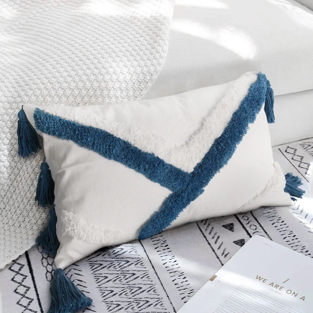 Coussin décoratif blanc et bleu avec des pompons et des motifs graphiques, posé sur un tapis de style ethnique.