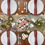 Une table de fête décorée avec des assiettes, des serviettes rouges, des œufs de Pâques colorés et des petits lapins blancs dans des nids, créant une ambiance printanière festive.