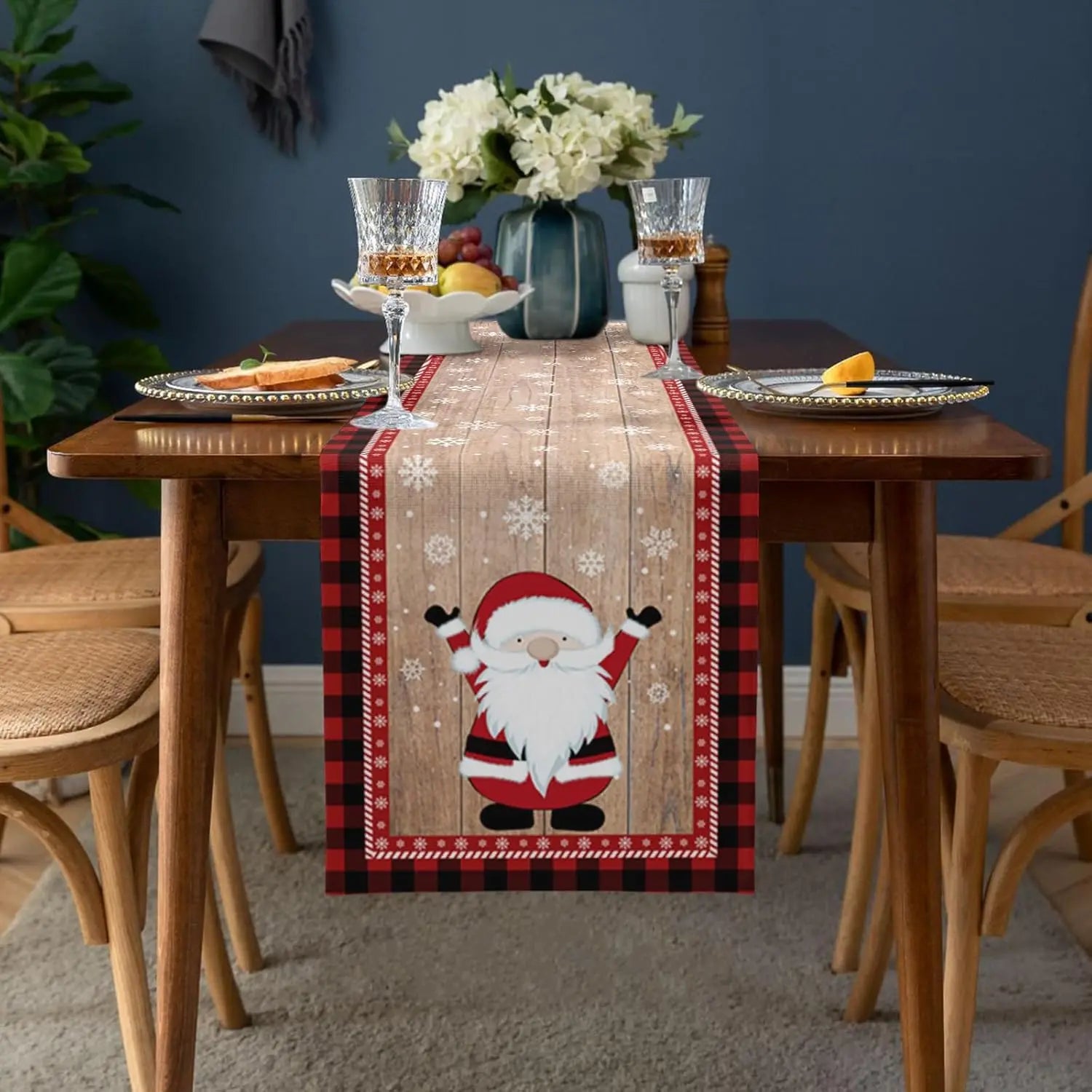 Cette image montre une table joliment décorée pour les fêtes de fin d'année, avec des fleurs blanches, des verres à vin et des fruits. Un chemin de table avec un motif de Père Noël et de flocons de neige ajoute une touche festive à la décoration.