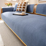 Un canapé bleu texturé avec un coussin décoratif en forme de boules brunes, posé sur un tapis gris clair dans un salon lumineux, avec un bouquet de fleurs sur une table basse.