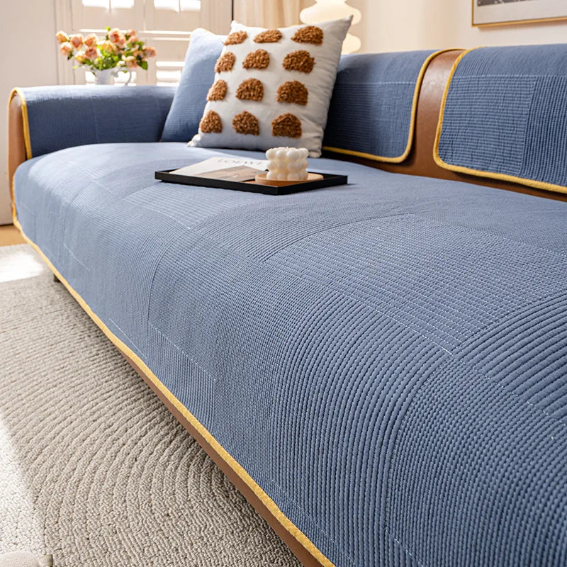 Un canapé bleu texturé avec un coussin décoratif en forme de boules brunes, posé sur un tapis gris clair dans un salon lumineux, avec un bouquet de fleurs sur une table basse.