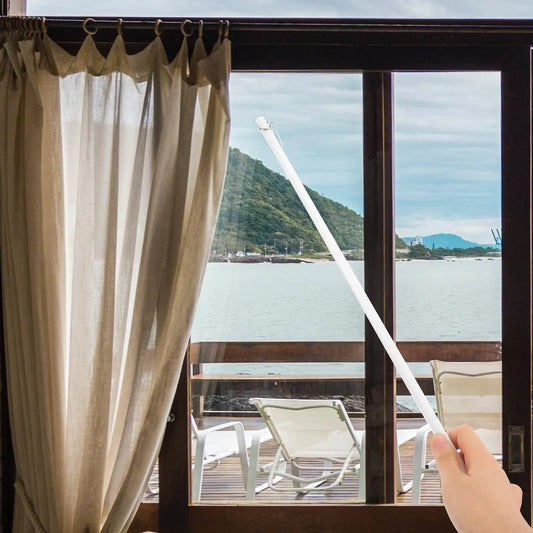 Une main utilise une baguette de tirage blanche pour ouvrir un rideau beige devant une baie vitrée avec vue sur la mer et les montagnes.