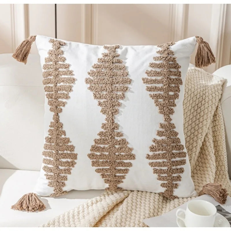 Trois oreillers décoratifs blancs avec des motifs graphiques beiges et des pompons assortis, sur un lit avec une couverture en tricot.