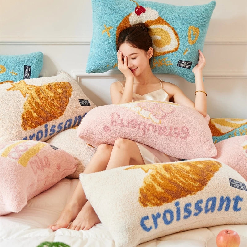 Une jeune femme souriante profite d'un moment de détente sur un lit confortable décoré de coussins aux motifs de viennoiseries.