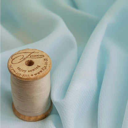Détail d’un voilage bleu clair en tissu léger accompagné d’une bobine de fil beige, illustrant la texture fluide et douce du tissu.