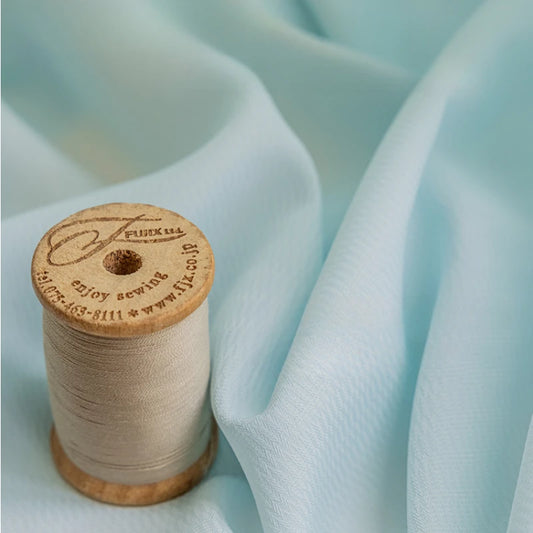 Détail d’un voilage bleu clair en tissu léger accompagné d’une bobine de fil beige, illustrant la texture fluide et douce du tissu.