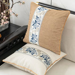 Deux coussins décoratifs en tissu beige et motifs bleus et gris ornent un canapé, accompagnés d'une branche fleurie dans un vase.